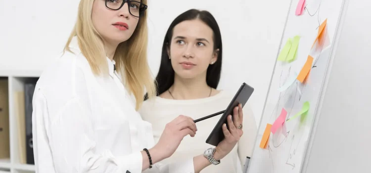 Două colege folosesc o tabletă lângă o tablă de prezentare